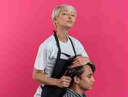 Foto gratuita giovane e bella barbiere femminile sicura in uniforme che tiene gli strumenti del barbiere e che fa taglio di capelli per il ragazzo isolato sulla parete rosa