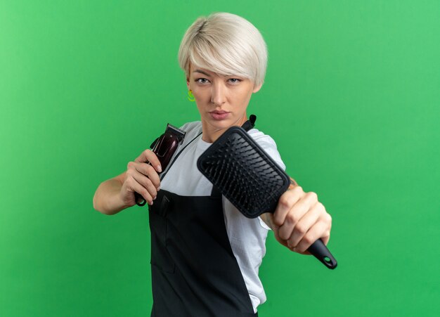 Confident young beautiful female barber in uniform holding out hair clipper with comb isolated on green wall