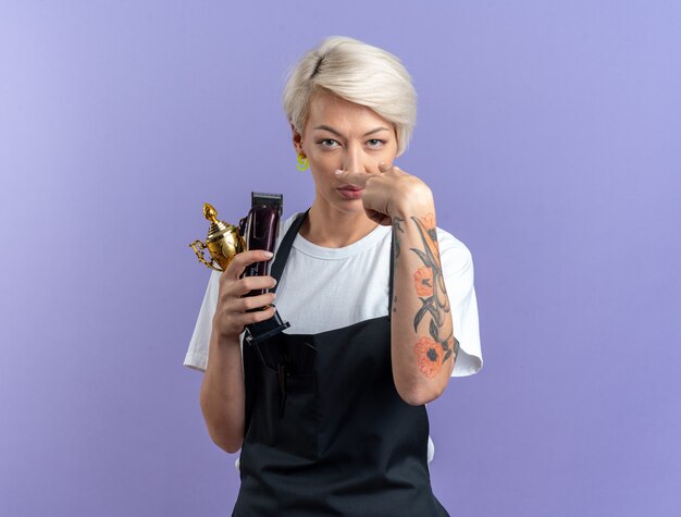 Free photo confident young beautiful female barber in uniform holding winner cup with hair clippers showing look gesture isolated on blue wall