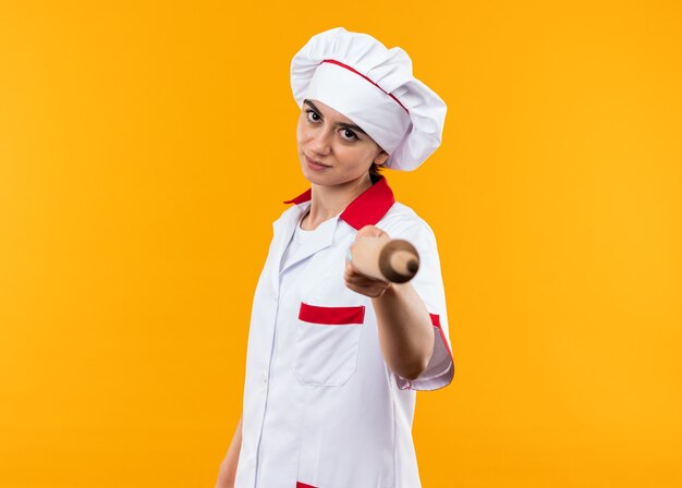 Confident young beautiful girl in chef uniform holding out rolling pin at camera 
