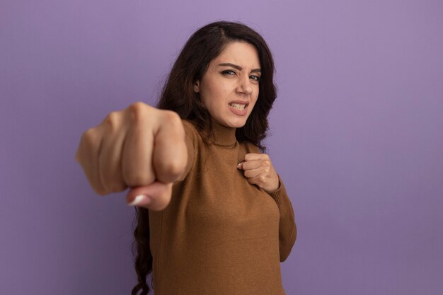 Confident young beautiful girl wearing brown turtleneck sweater holding out fist isolated on purple wall with copy space