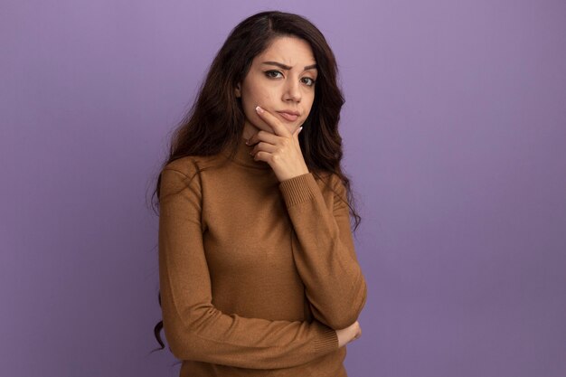 Confident young beautiful girl wearing brown turtleneck sweater putting hand on chin isolated on purple wall with copy space