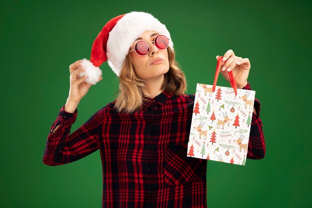Confident young beautiful girl wearing christmas hat with glasses holding gift bag isolated on green background