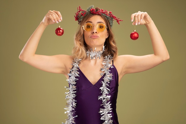 無料写真 オリーブグリーンの背景で隔離のクリスマスツリーのボールを保持している首に花輪と花輪と紫色のドレスとメガネを身に着けている自信を持って若い美しい少女