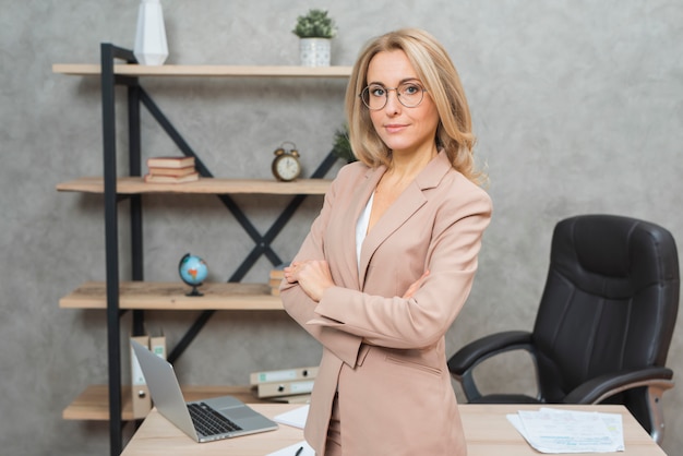 Confident young blonde businesswoman standing in front of office desk