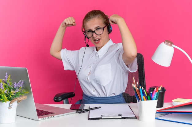 Confident young blonde call centre girl wearing headset and glasses sitting at desk with work tools looking at camera doing strong gesture isolated on pink wall