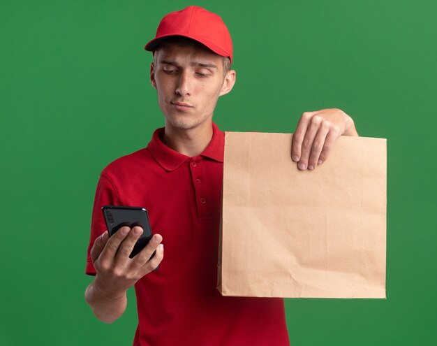 Уверенный молодой блондин доставки мальчик держит бумажный пакет и смотрит на телефон