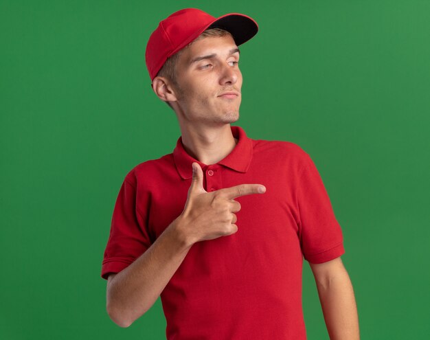 Free photo confident young blonde delivery boy looks and points at side on green