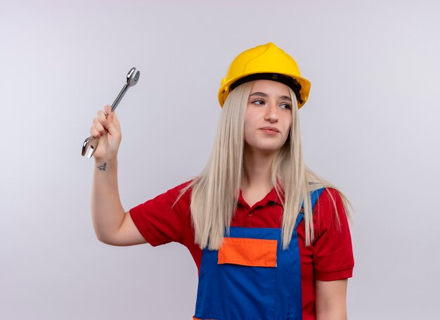 Foto gratuita ragazza bionda giovane fiduciosa del costruttore dell'ingegnere in uniforme che alza la chiave aperta che esamina il lato destro sullo spazio bianco isolato