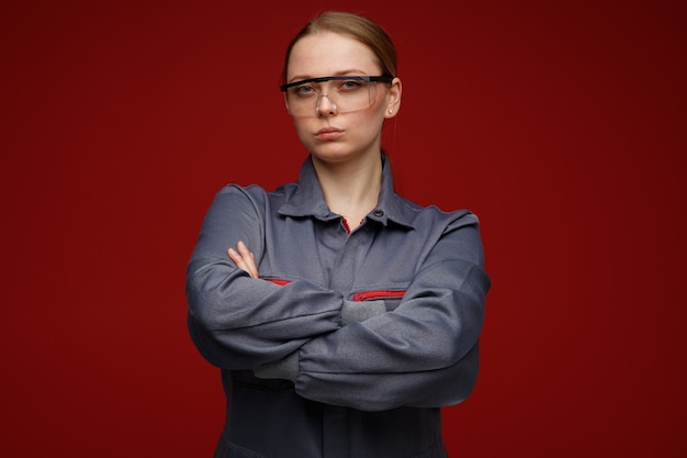 Confident young blonde female engineer wearing uniform and safety glasses standing with closed posture 