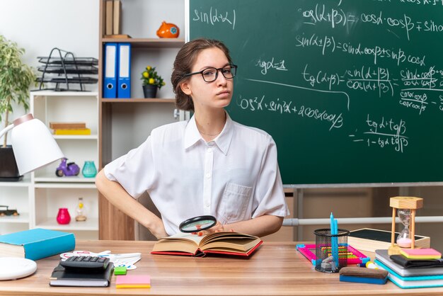 Confident young blonde female math teacher wearing glasses sitting at desk with school tools  keeping hand on waist and on open book with magnifying glass in hand in classroom