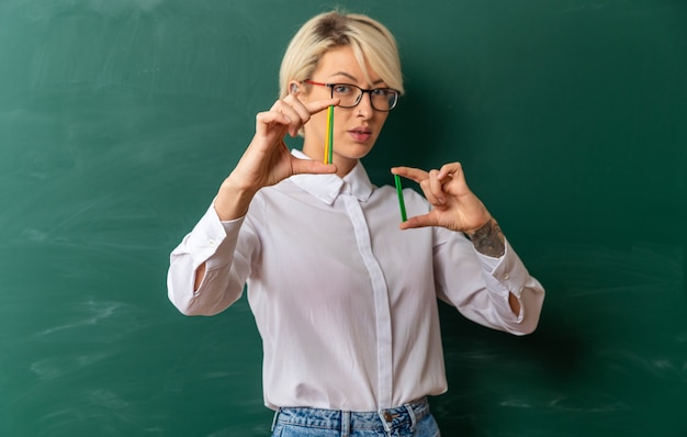 Free photo confident young blonde female teacher wearing glasses in classroom standing in front of chalkboard showing counting sticks looking at camera