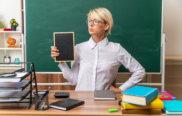 confident young blonde female teacher wearing glasses sitting at desk with school supplies in classroom showing mini blackboard looking at it keeping hand on waist