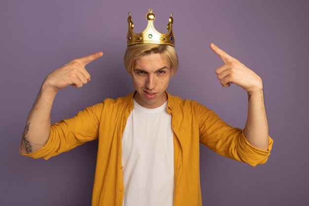 Free photo confident young blonde guy wearing yellow t-shirt and crown points at himself isolated on purple