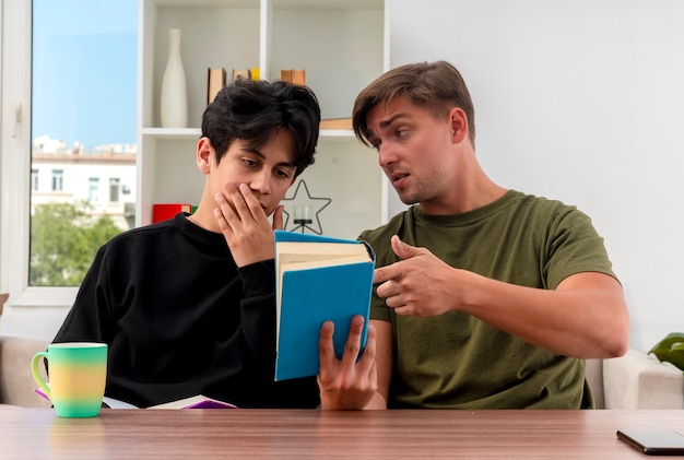Confident young blonde handsome man holding and pointing at book sitting at table and looking at young brunette handsome guy putting hand on mouth looking at book inside living room