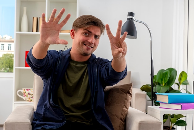 Free photo confident young blonde handsome man sits on armchair gesturing eight with fingers inside living room