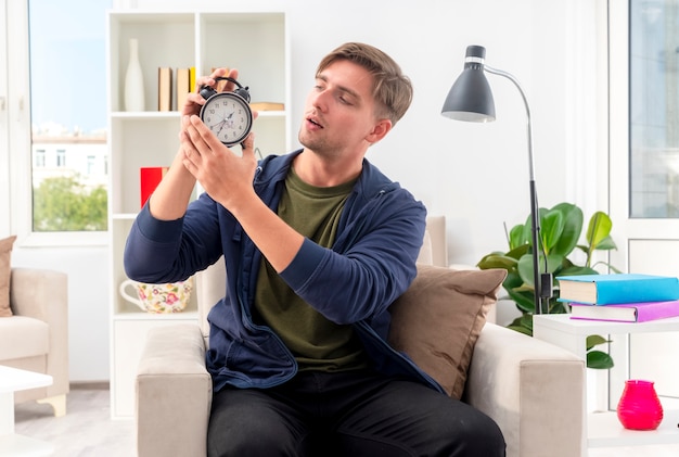 Confident young blonde handsome man sits on armchair holding and looking at alarm clock inside the living room