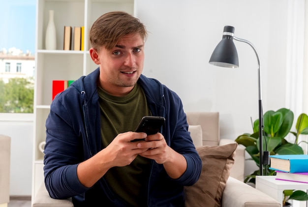 Confident young blonde handsome man sits on armchair holding phone