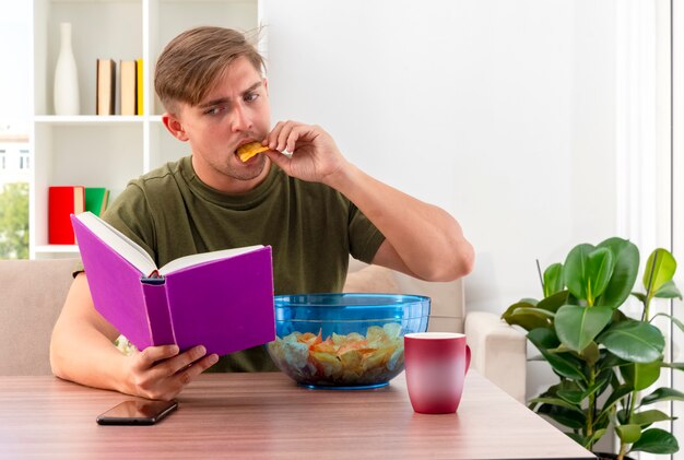 Confident young blonde handsome man sits at table with bowl of chips cup and phone holding and looking at book eating chips inside living room