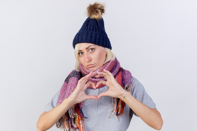 Free photo confident young blonde ill slavic woman wearing winter hat and scarf gestures heart hand sign isolated on white wall with copy space