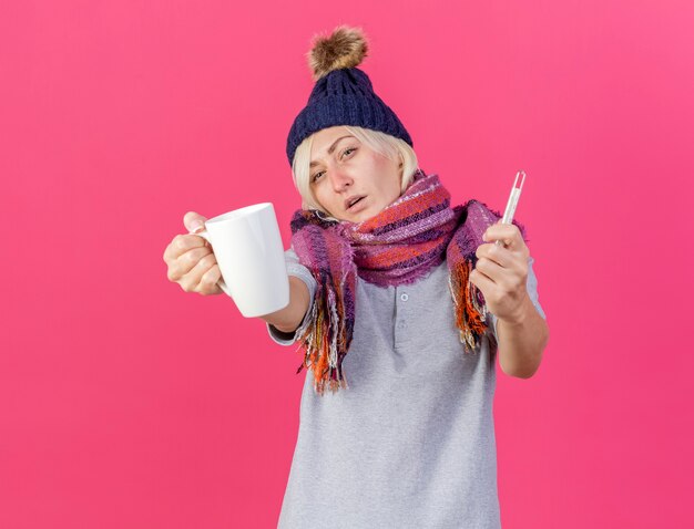 冬の帽子とスカーフを身に着けている自信を持って若い金髪の病気のスラブ女性は、コピースペースでピンクの壁に分離されたカップと温度計を保持します