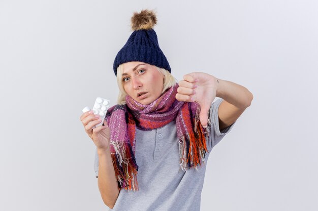 Confident young blonde ill slavic woman wearing winter hat and scarf thumbs down and holds holds medicine in glass bottle with pack of medical pills isolated on white wall with copy space