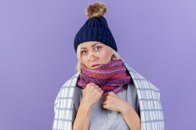 Confident young blonde ill slavic woman with medical plaster on nose wearing winter hat and scarf