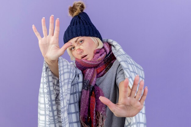 Confident young blonde ill slavic woman with medical plaster on nose wearing winter hat and scarf