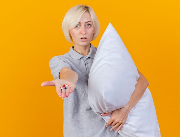 Confident young blonde ill woman hugs pillow and holds medical pills in hand isolated on orange wall