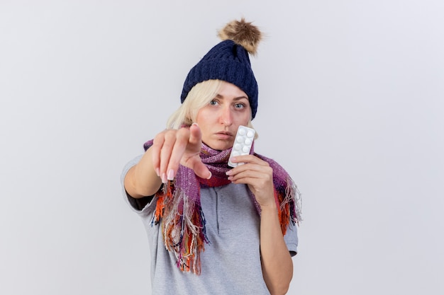 Confident young blonde ill woman wearing winter hat and scarf holds pack of medical pills pointing at front isolated on white wall