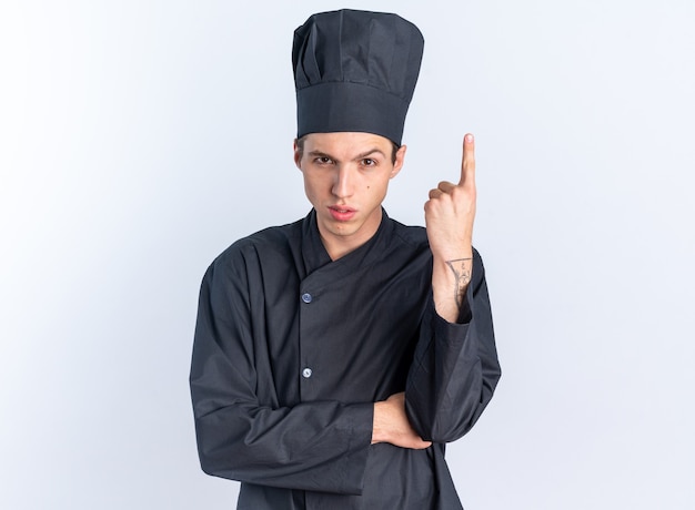 Confident young blonde male cook in chef uniform and cap looking at camera looking at camera pointing up isolated on white wall with copy space