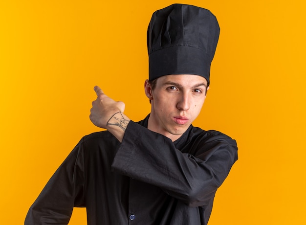 Confident young blonde male cook in chef uniform and cap looking at camera pointing behind isolated on orange wall