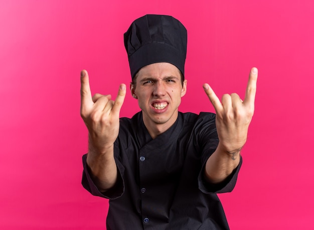 Confident young blonde male cook in chef uniform and cap looking at camera showing teeth doing rock sign isolated on pink wall