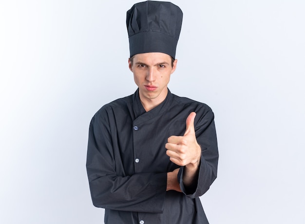 Confident young blonde male cook in chef uniform and cap looking at camera showing thumb up isolated on white wall with copy space