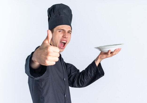 Confident young blonde male cook in chef uniform and cap standing in profile view stretching out plate  stretching out hand towards showing thumb up screaming isolated