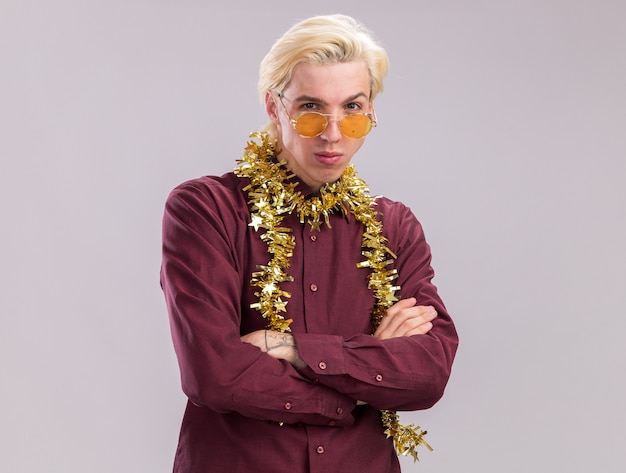 Confident young blonde man wearing glasses with tinsel garland around neck standing with closed posture looking at camera isolated on white background with copy space