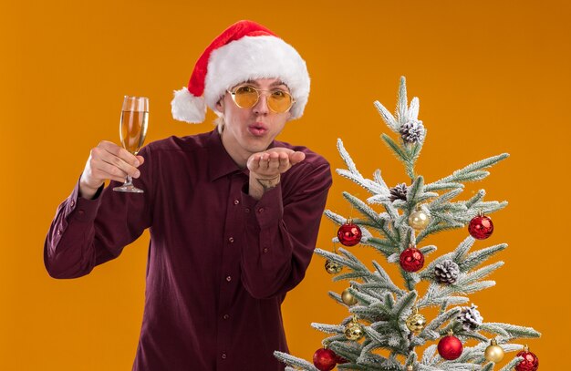 Confident young blonde man wearing santa hat and glasses standing near decorated christmas tree holding glass of champagne looking at camera sending blow kiss isolated on orange background