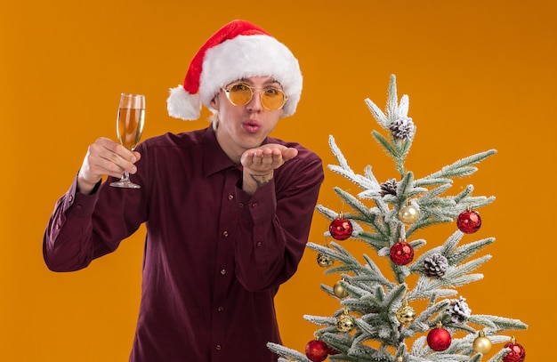 오렌지 배경에 고립 타격 키스를 보내는 카메라를보고 샴페인 잔을 들고 장식 된 크리스마스 트리 근처에 서있는 산타 모자와 안경을 착용 확신 젊은 금발의 남자