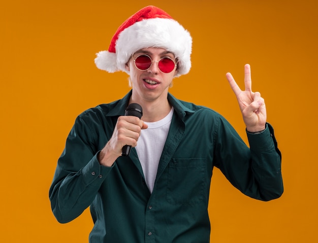 산타 모자와 안경을 쓰고 자신감이 젊은 금발의 남자가 오렌지 배경에 고립 된 평화 서명을하고 카메라를보고 마이크에 이야기