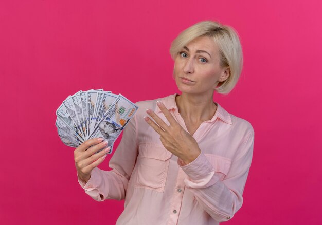 Уверенная молодая блондинка славянская женщина, держащая деньги и показывающая трое рукой, изолированной на розовом фоне
