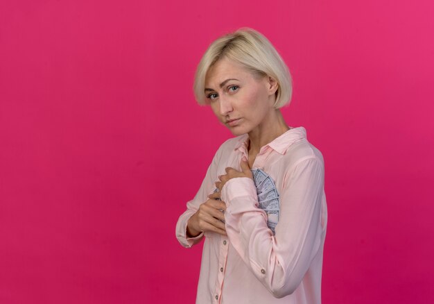 Confident young blonde slavic woman standing in profile view holding money isolated on pink background with copy space