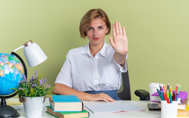 Confident young blonde student girl sitting at desk with school tools doing stop gesture 
