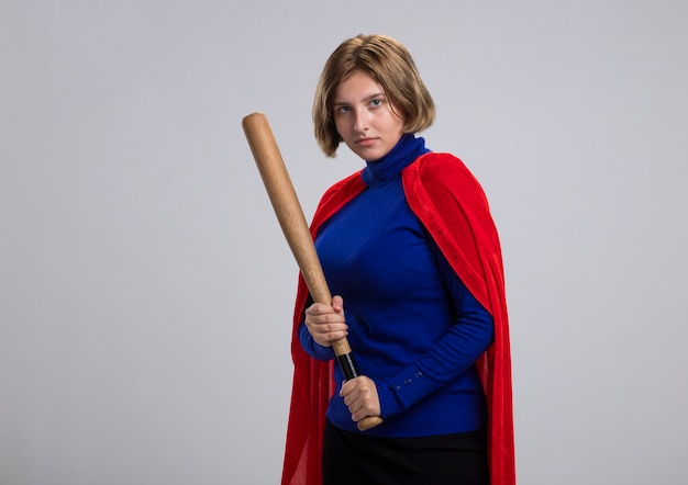 Confident young blonde superhero girl in red cape standing in profile view holding baseball bat  isolated on white wall with copy space