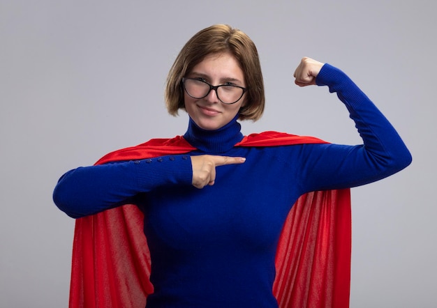 Confident young blonde superhero woman in red cape wearing glasses doing strong gesture looking at front pointing at her muscles isolated on white wall