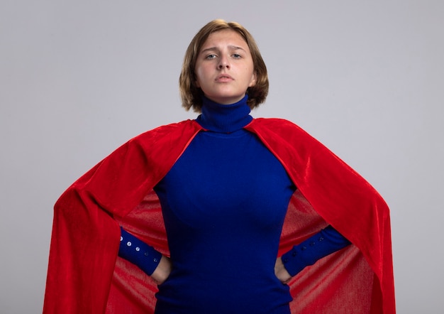 Confident young blonde superwoman in red cape keeping hands on waist looking at front isolated on white wall