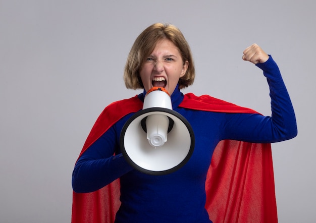 Confident young blonde superwoman in red cape looking at front doing strong gesture shouting in loud speaker isolated on white wall