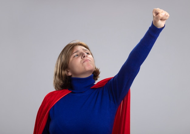 Free photo confident young blonde superwoman in red cape raising fist up looking at it isolated on white wall