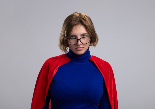 Confident young blonde superwoman in red cape wearing glasses looking at front isolated on white wall