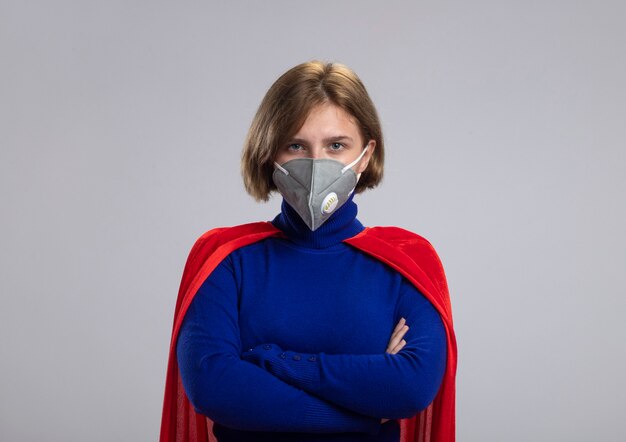 Confident young blonde superwoman in red cape wearing protective mask standing with closed posture looking at front isolated on white wall