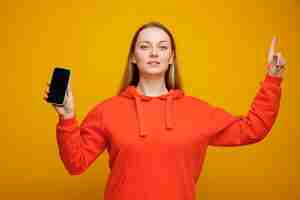 Free photo confident young blonde woman holding mobile phone pointing up